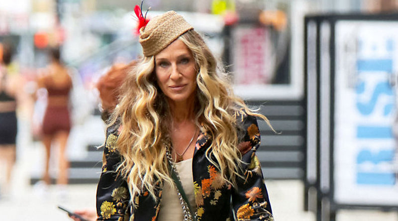 A woman with long, voluminous blonde wavy hair walking outdoors in an urban setting with blurred signage in the background. Featured image for blog post: And Just Like That, How to Create the TV Show-Inspired Curls