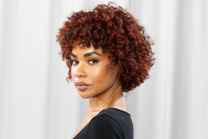 A woman with voluminous, tightly coiled curly hair in a rich dimensional copper-red tone gazes over her shoulder wearing a black top, photographed against a light grey draped background. Featured image for blog post: Achieving Dimensional Copper Hair for 