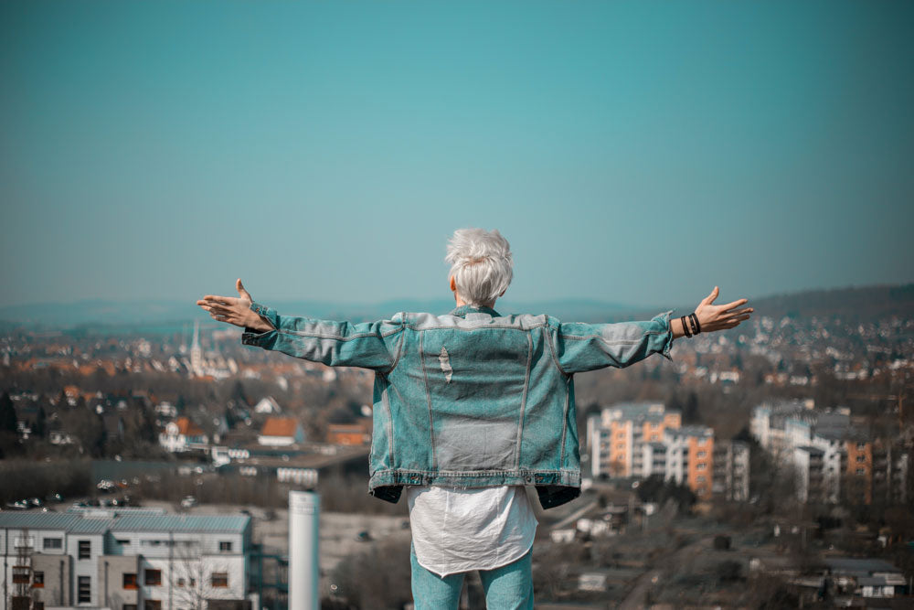 A person with a short, silvery-white pixie cut is seen from behind, arms outstretched wide, wearing a denim jacket while standing at a scenic overlook above a cityscape. Featured image for blog post: GRAY IS ANATOMY 