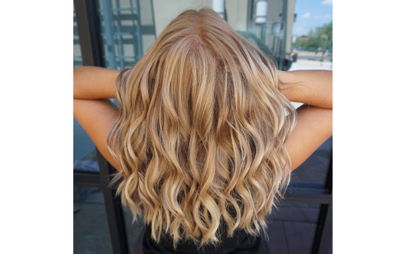 Back view of a woman with medium-length, beachy wavy champagne blonde hair with warm golden highlights, her arms raised behind her head against an urban outdoor backdrop. Featured image for blog post:  Set The Tone for NYE with Bubbly & Champagne Blonde H