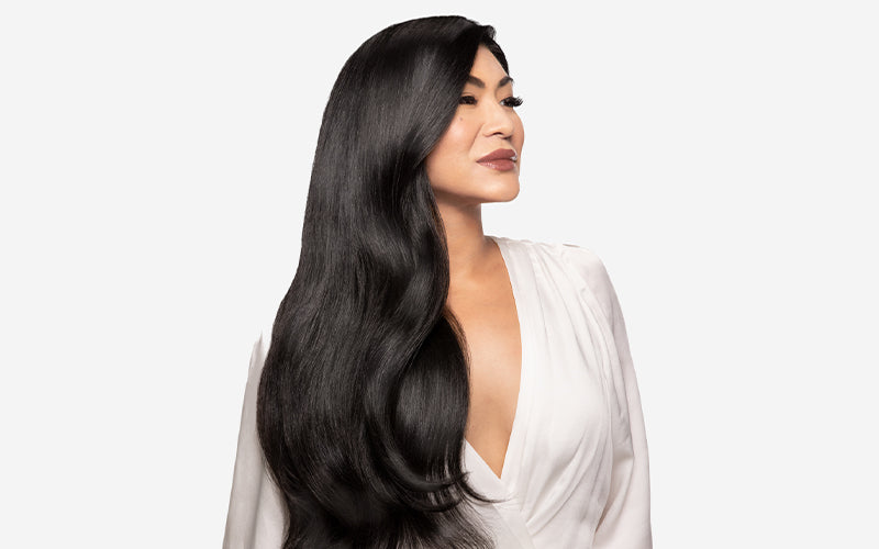 A woman with long, silky jet-black hair styled in sweeping, voluminous waves looks to the side wearing a white draped blouse, photographed against a clean white background. Featured image for blog post: Step by Step Sleek Blow-Dry using Pearl Detangler