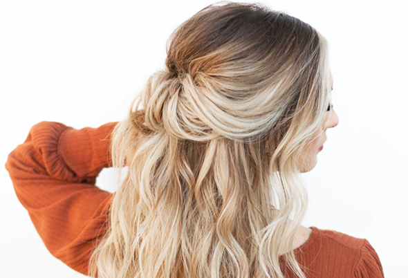 A woman seen from behind with long, wavy blonde hair pulled into a loose half-updo, wearing a rust-orange sweater against a soft light background. Featured image for blog post: Holiday Hair Hacks with Stephanie Brinkerhoff