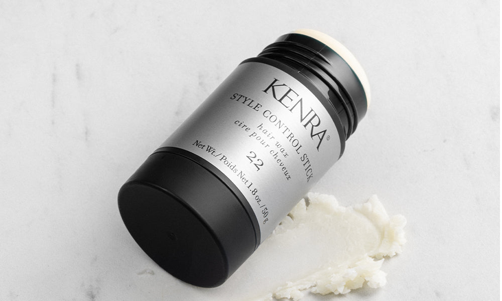 The Kenra Style Control Stick 22 hair wax product lying on its side on a white marble surface, with a small amount of white waxy product scooped out beside it, showing its matte black and silver cylindrical packaging. Featured image for blog post: Introdu