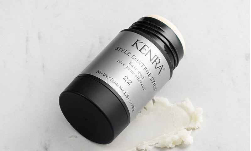 The Kenra Style Control Stick 22 hair wax product lying on its side on a white marble surface, with a small amount of white waxy product scooped out beside it, showing its matte black and silver cylindrical packaging. Featured image for blog post: Introdu