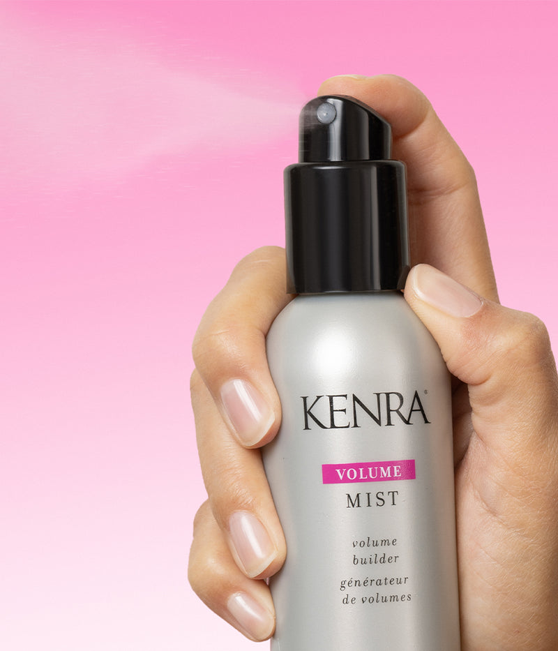 A hand holding a Kenra Volume Mist spray bottle — silver with a pink label — with a fine mist spraying from the nozzle against a soft pink gradient background. Featured image for blog post: How to Get Extra Volume in Your Hair with Volume Mist 2