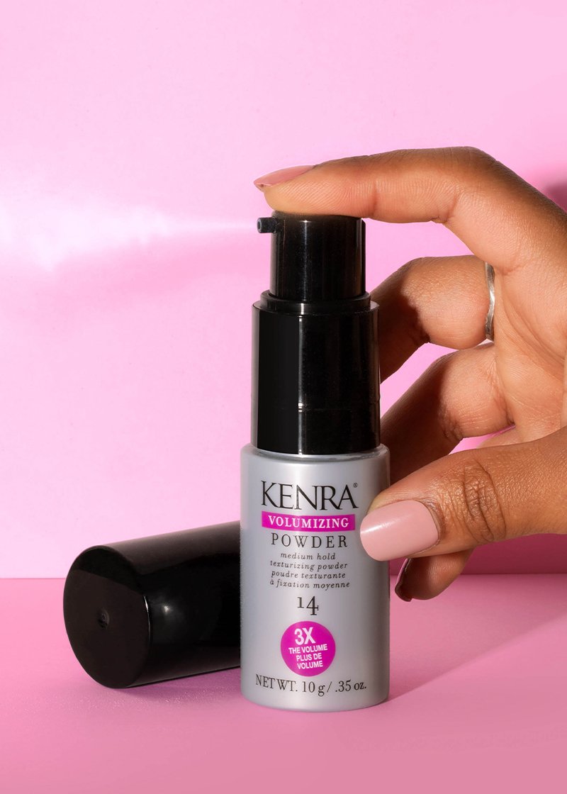 A hand presses the pump of a Kenra Volumizing Powder 14 bottle against a bright pink background, with another capped bottle resting on its side below. Featured image for blog post: Top 5 Stocking Stuffers for the Holiday Season!