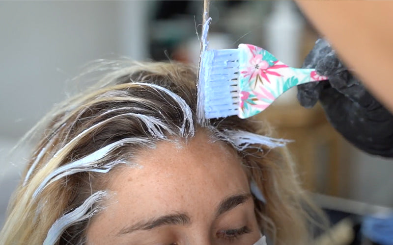Close-up of a colorist applying foil babylights to a blonde woman’s hair, with fine sections of hair coated in lightener fanned out across the scalp. Featured image for blog post: Everything You Need To Know About Babylights