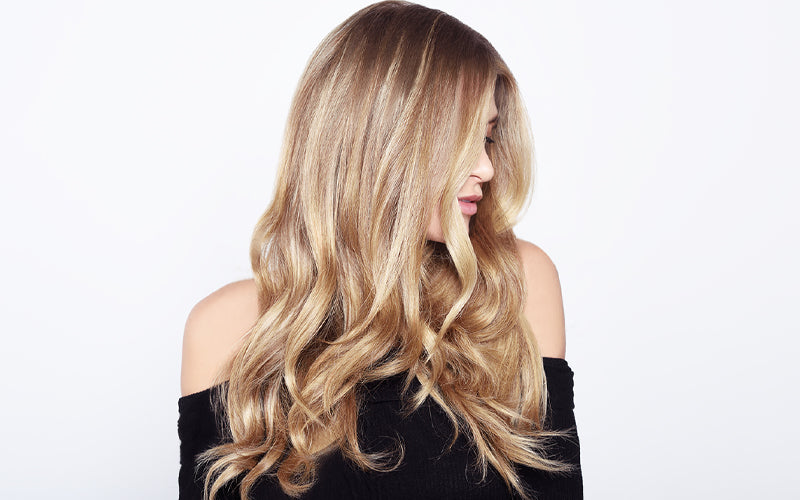 A woman facing away with long, voluminous blonde hair featuring seamless balayage highlights blending from darker roots to bright golden ends, styled in loose, flowing waves. Featured image for blog post: Ombré vs. Balayage: Which Style Is Right for You?