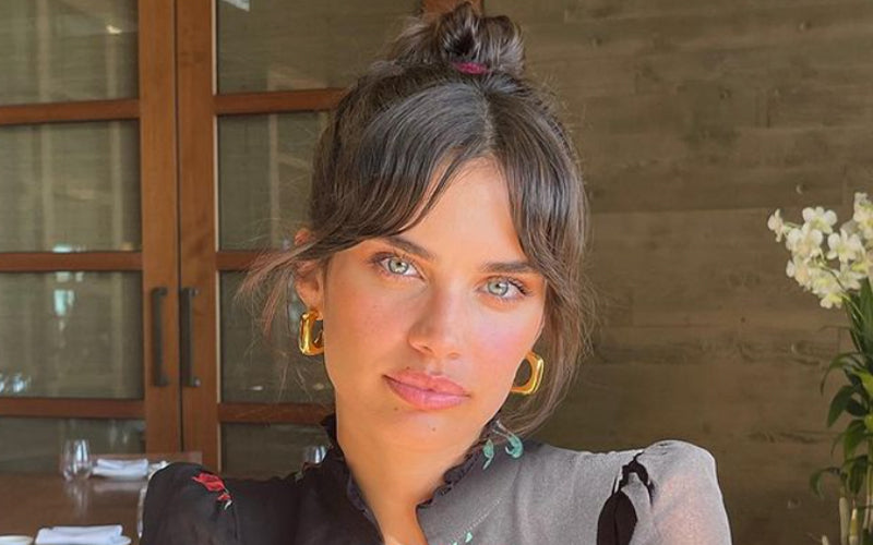 A woman with dark, center-parted hair styled into a sleek bun faces forward, showing neatly groomed eyebrow-grazing micro bangs framing her face. Featured image for blog post: Bangs are Back!