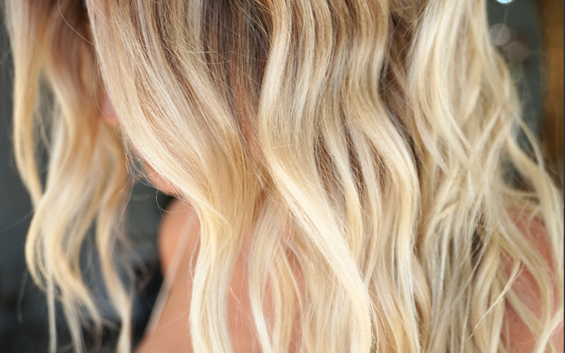 Back view of long, flowing hair with warm golden-to-platinum blonde balayage, the strands blown out smooth and glossy with visible dimension. Featured image for blog post: How to Get Beach Blonde Hair Year-Round