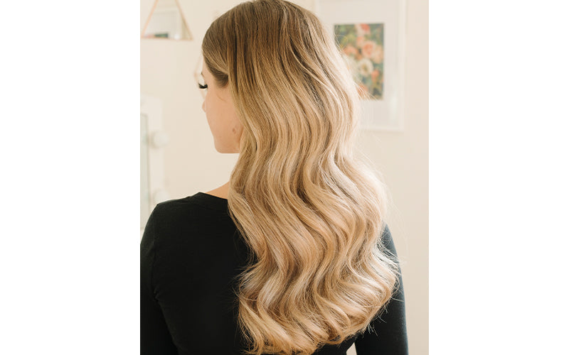 A woman photographed from behind showing long, lush blonde hair styled in large, loose waves with a warm honey-to-ash gradient, against a soft blurred background. Featured image for blog post: Big Waves for Big Holidays
