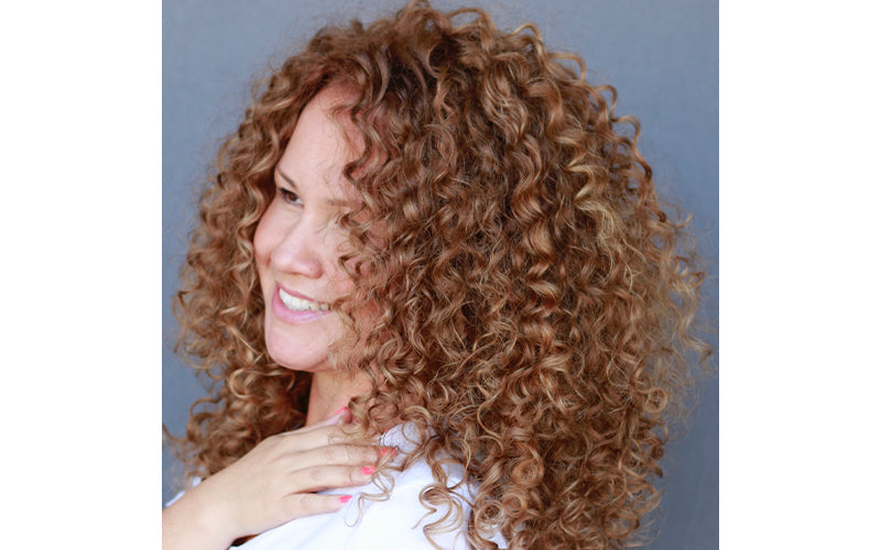 A profile portrait of a woman with a full head of voluminous, defined auburn-brown curls cascading past her shoulders, smiling softly against a light neutral background. Featured image for blog post: How To Sleep With Curly Hair Without Ruining It