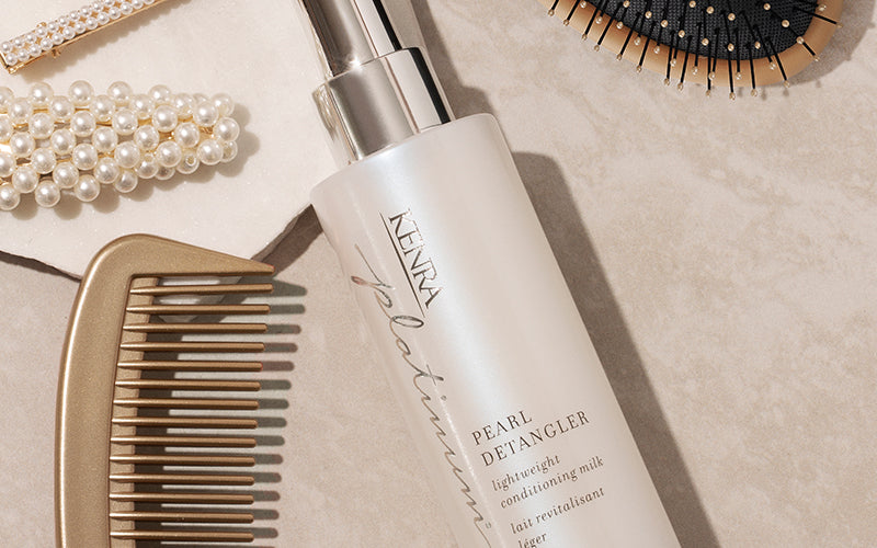 A wooden wide-tooth comb and a bottle of hair detangling spray resting on a neutral surface alongside a wooden hairbrush and other haircare accessories. Featured image for blog post: How To Detangle Hair Without Causing Damage