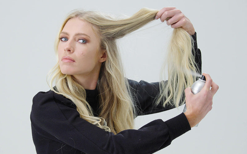 Close-up of a person lifting sections of long, straight blonde hair while spraying dry shampoo directly at the roots, against a light grey background. Featured image for blog post: A Step-by-Step Guide on How Dry Shampoo Works