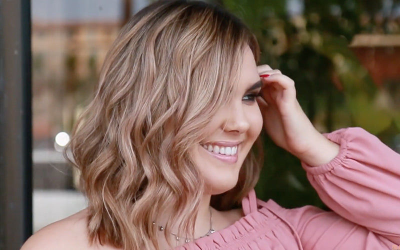 A smiling woman in a pink blouse with soft, wavy blonde hair photographed outdoors in warm natural light, looking off to the side. Featured image for blog post: Best of Kenra Color Formulas