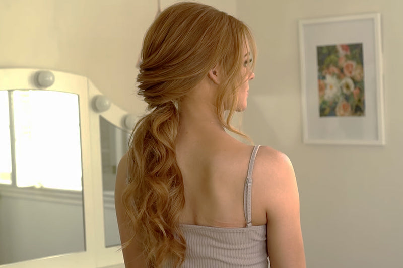 A styled low ponytail with a braided or twisted rope-like section running down the back is reflected in a vanity mirror with lit bulbs, showing the intricate texture of the finished look. Featured image for blog post: Step by Step: How to Create a Low, Fr