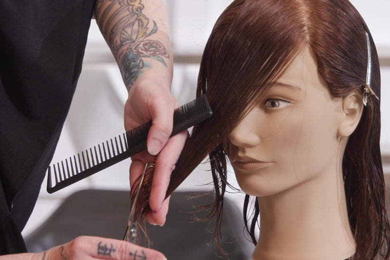 A tattooed stylist's hands use a comb and scissors to trim a client's dark hair in a close-up salon shot. Featured image for blog post: Fringe 101: 4 Different Ways to Cut Bangs