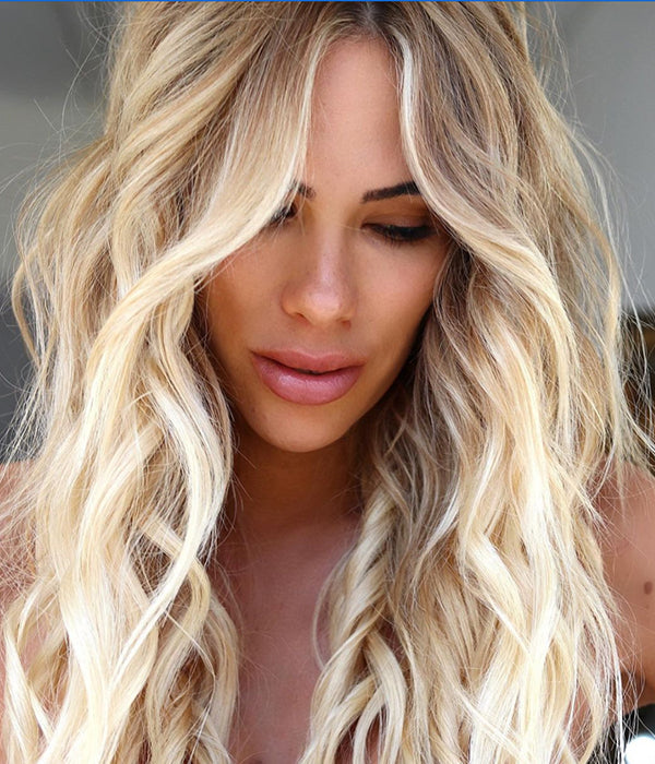 A woman with long, voluminous wavy platinum blonde hair faces the camera against a bright, sun-drenched background, showcasing a rich golden-blonde color result. Featured image for blog post: Get Golden with the Kenra Color Sunrise Collection 