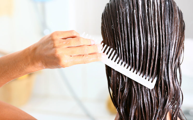 Close-up of wet dark hair being combed through with a wide-tooth comb in the shower, with conditioning product visibly coating the strands. Featured image for blog post: How To Use Leave-In Conditioner for Healthy Hair