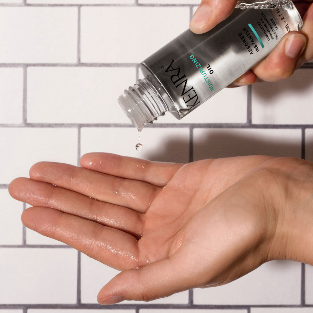 Kenra Moisturizing Oil bottle dispensing a drop of oil onto an open palm against a white subway tile background