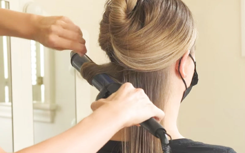 A close-up of a stylist's hands using a round brush and blow dryer on a section of medium brown hair, with the hair pulled taut and slightly frizzy texture visible, suggesting heat styling in progress. Featured image for blog post: Can Heat Damaged Hair B