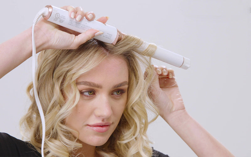 A woman with curly, light blonde hair uses a curling wand on a section of her hair, holding the barrel close to her face against a soft white background. Featured image for blog post: Does Heat Protectant Actually Work and Do You Need It?