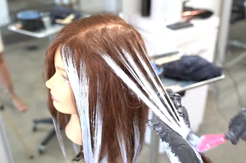 Close-up of a balayage technique in progress, with long streaks of lightener painted onto sections of brown hair pulled away from the head. Featured image for blog post: How to Do a High-Speed Balayage