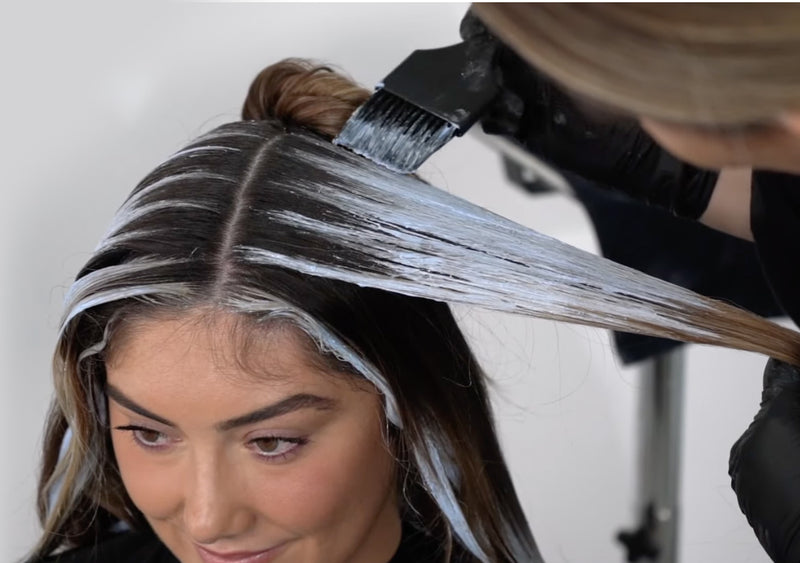 A stylist applies white balayage lightener to sections of a client's dark brown hair using a brush, with foil sectioning visible in this overhead in-salon process shot. Featured image for blog post: How to Create High Point Balayage on Grown-Out Hair