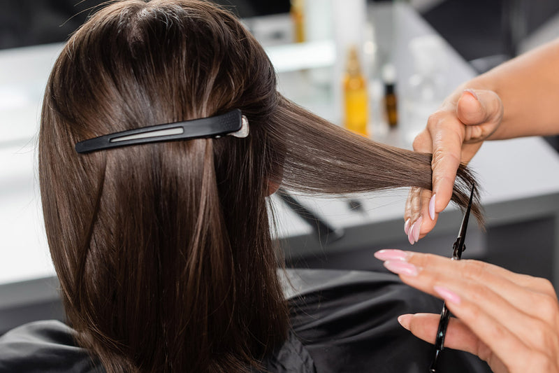 A close-up of a stylist's hands using scissors to cut a section of long, straight dark brunette hair at a salon, demonstrating a precision layering technique. Featured image for blog post: Get Gorgeous Hair with Ghost Layers: The Ultimate Invisible Layeri