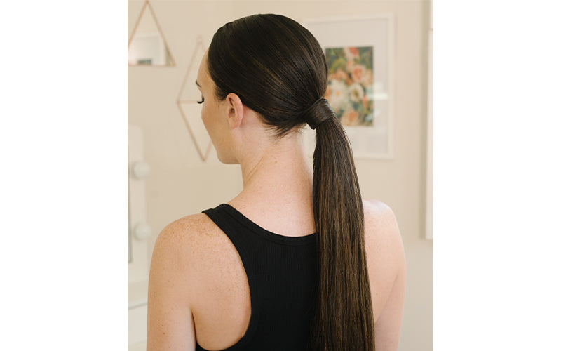 Back view of a woman with long sleek dark hair styled in a low ponytail, wearing a black tank top