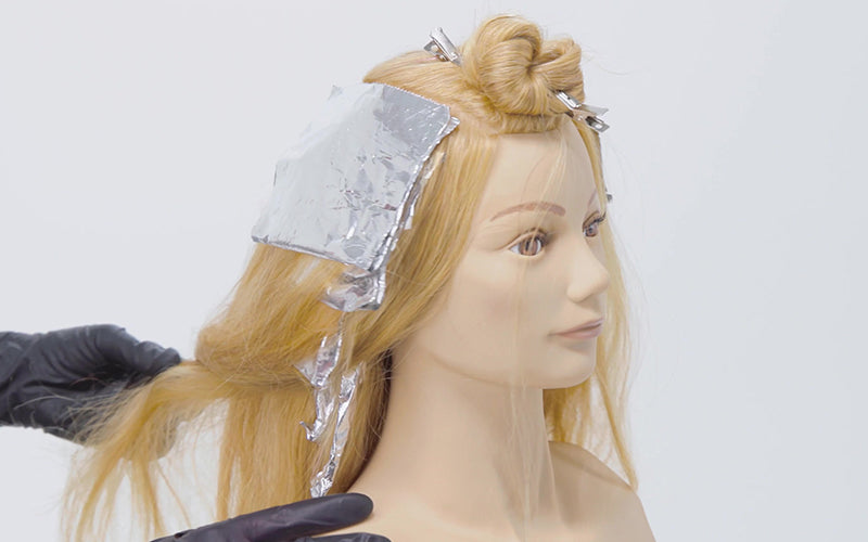 A close-up of a blonde mannequin head with silver foils being applied to sectioned hair during a coloring service, illustrating a highlights or lowlights technique in progress. Featured image for blog post: Highlights vs. Lowlights: Which Color is Best fo