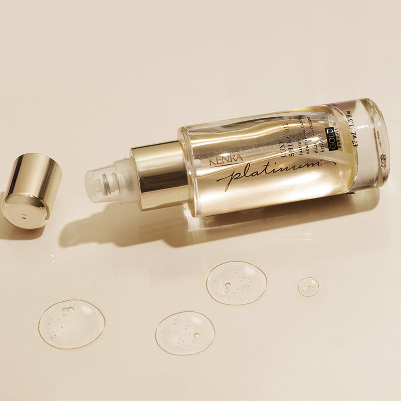 A close-up product shot of a slender, glass-like Kenra LuxeOil treatment bottle lying on its side against a neutral light background. Featured image for blog post: HELP YOUR HAIR SHINE LIKE GOLD