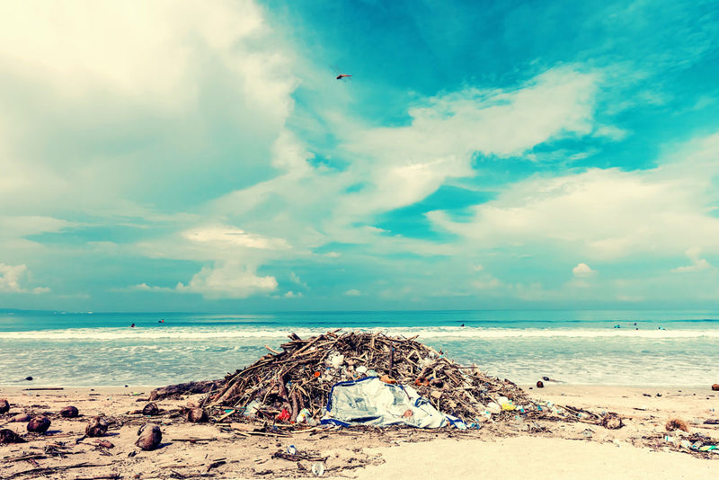 A large pile of plastic waste and debris sits on a beach under a bright blue sky with scattered clouds, evoking ocean pollution and the need for eco-conscious choices. Featured image for blog post: BECOME MORE EC0-FRIENDLY