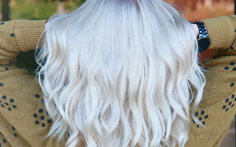 A close-up of a model with bright white-platinum silver wavy hair, softly styled and photographed outdoors with a blurred natural background. Featured image for blog post: How To Get Silver Hair: Everything You Need To Know
