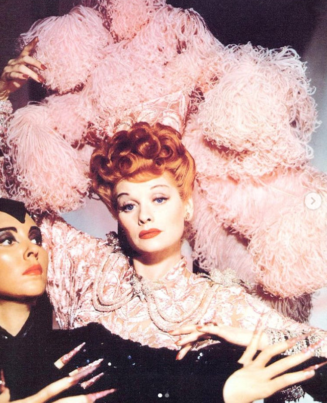 A woman with voluminous bright red hair posed dramatically alongside a male companion against a backdrop of large pink feathery flowers, evoking vintage theatrical glamour. Featured image for blog post: Redheads Throughout the Decades 