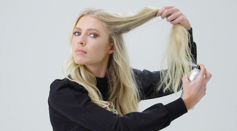 A woman holds and lightly mists a section of her long, straight blonde hair while working styling product through it, against a clean white background. Featured image for blog post: Tips & Tricks on How to Get the Best Second (or Third) Day Hair