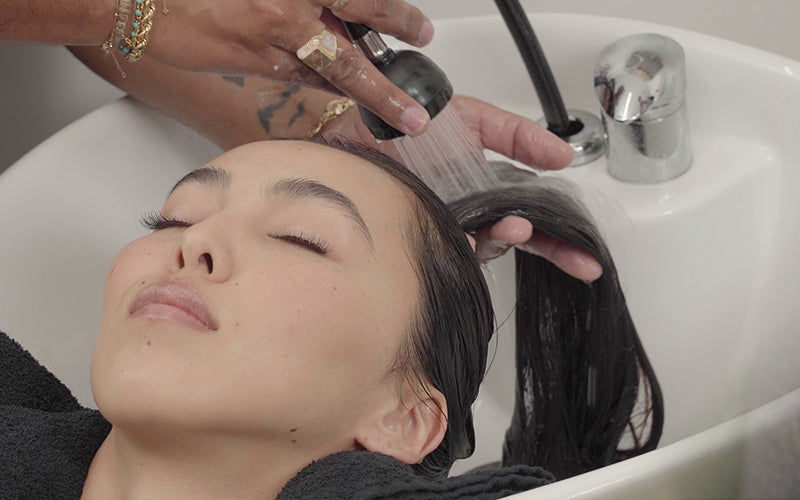 A person with dark hair leaning back at a salon shampoo bowl while a stylist rinses their hair under running water. Featured image for blog post: Here Is Our Top Shampoo For Oily Hair, Plus a Few More