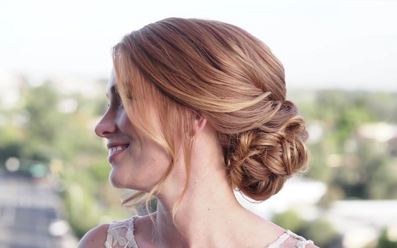 A woman’s hair is styled into an elegant low chignon or twisted updo, with soft face-framing pieces, photographed from behind against a softly blurred outdoor backdrop. Featured image for blog post: Simple Time-saving Hairstyles for Holiday Festivities