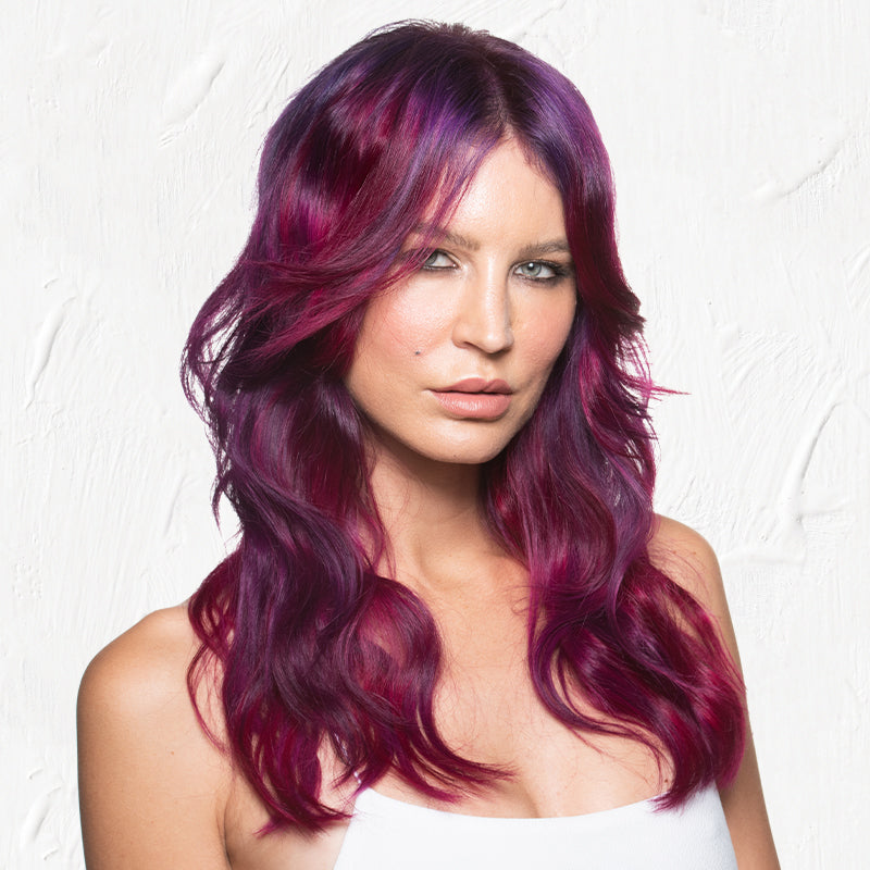 A woman with vibrant magenta-to-violet wavy hair, styled in loose waves falling past her shoulders with rich jewel-toned color blending from pink to deep purple. Featured image for blog post: Cotton Candy Tri-Color Melt Using Kenra Creatives