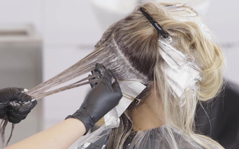 A stylist wearing black gloves applying hair color with a brush to sections of light brown hair that are clipped and parted for foiling. Featured image for blog post: How To Go From Brunette to Blonde Effortlessly