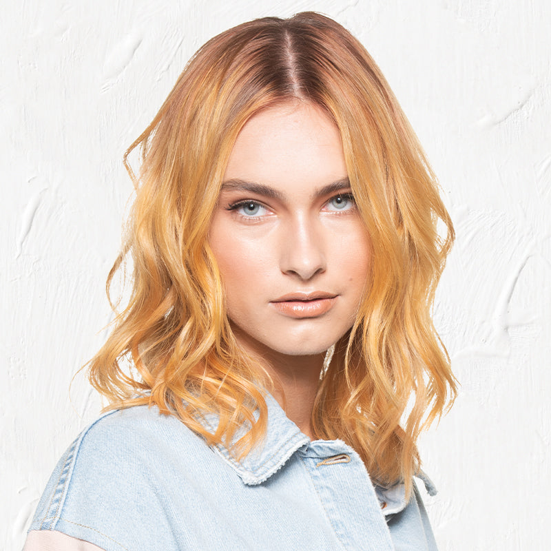 A close-up portrait of a woman with a warm golden-copper color melt, with soft wavy texture and sun-kissed peachy-blonde tones blending from roots to ends, against a neutral background. Featured image for blog post: Warm Golden Color Melt Using Kenra Crea